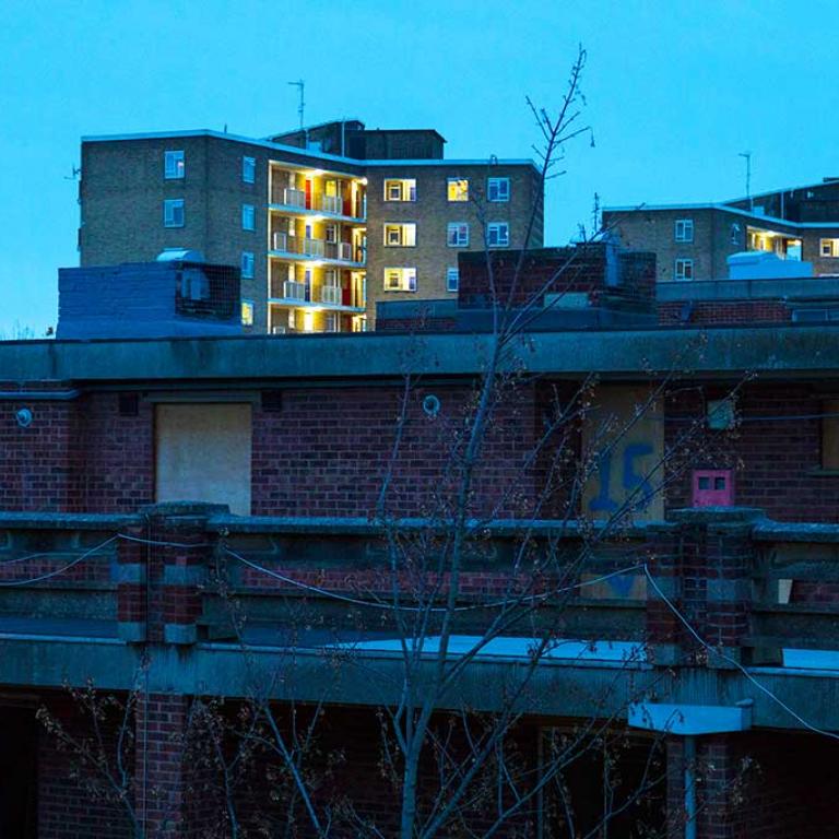 The top floor flats at Murchison House with the doors of number 14 and 15 boarded up prior to demolition. In the background, Treverton Tower is lit up at nightfall. Photo taken by Patrick Gorman, 2019.