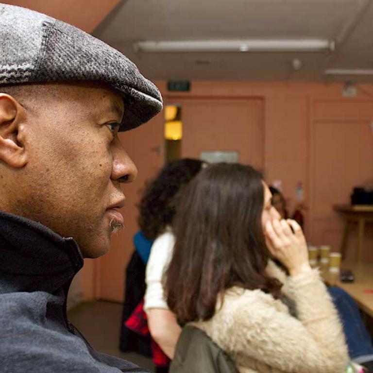 Junior Tomlin, local resident and artist, at the Photography Collation Workshop. His profile, including his flat cap, is in the left foreground. To the right are the profiles of other participants sitting at a table taking part in the workshop run by Kevin Percival.