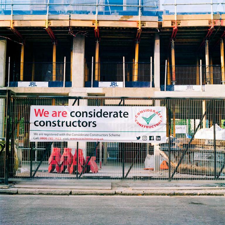 Construction Signage in front of a new block of flats called 'Bond Mansions', during construction on the site of Edward Kennedy House and Pepler House. The image shows a sign reading: ‘We Are Considerate Constructors’, on the gate of the building site, with the skeleton of the new building in the background. Photo by Kevin Percival, a resident of Pepler House, 2018.