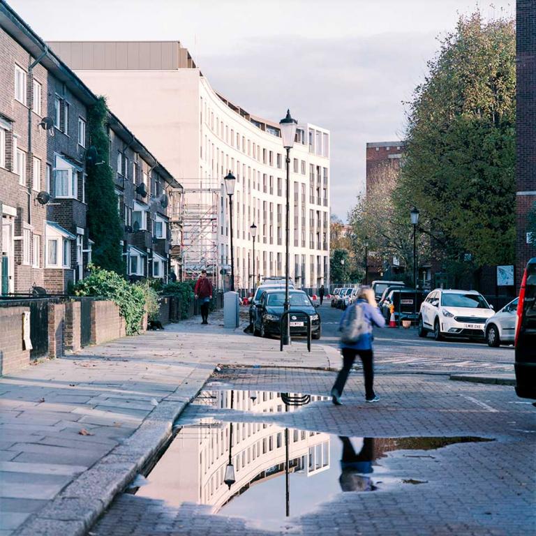 A view looking East along Wornington Road. From left to right: Pepler House, Bond Mansions, and Watts House. A woman crosses the street in the mid-ground and a large puddle in the foreground reflects Bond Mansions, lit up by strong evening sunlight. Photo by Kevin Percival, a resident of Pepler House, 2019.