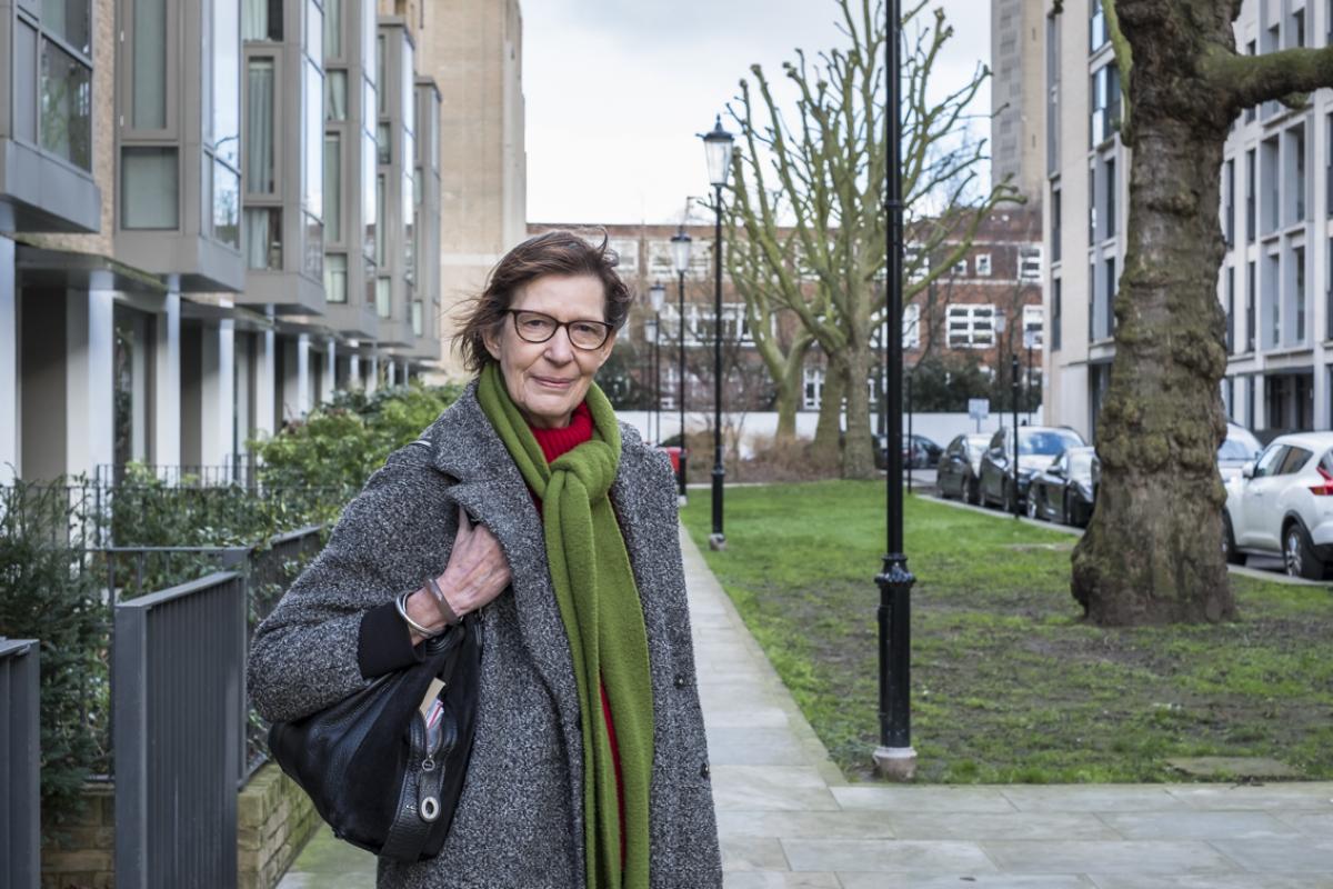 Eve is standing on the pavement. To the right of the pavement is a strip of grass with a large tree and lamp posts going down the side of the grass strip. On the right of the grass are parked cars. Eve has shortish brown hair and dark brown framed glasses. She is wearing a grey wool coat with a red polo neck jumper underneath and a green woollen scarf. She has a black handbag over her right shoulder and three bangles of her right wrist. The day is overcast.