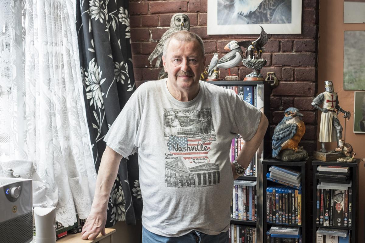 Keith is standing in his living room by the window which has grey and white flower patterned curtains with white viole curtains behind. Keith is wearing a Nashville t-shirt and behind him is a black bookcase filled with books and DVD's. There are ornaments of various sizes sitting atop the bookcase; 5 birds including an owl, a puffin and 2 eagles, a medeival knight in full armour and a small ornament of an otter.