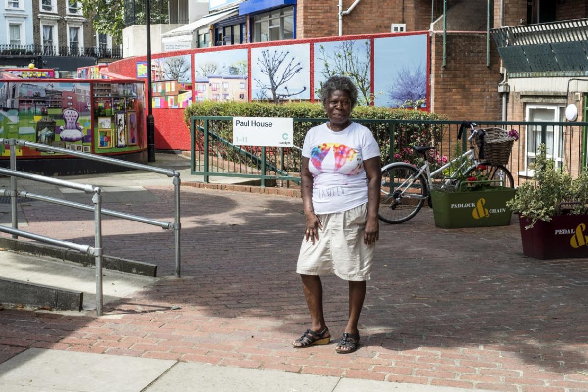 Cheryl is standing in the Wornington Green Estate in front of Paul House. In the background there is a brick wall covered in large images to the left, directly behind Cheryl are green railings, a bicycle and two plant pots. Cheryl is wearing a white t-shirt with a band of colour, cream coloured shorts and black sandals.