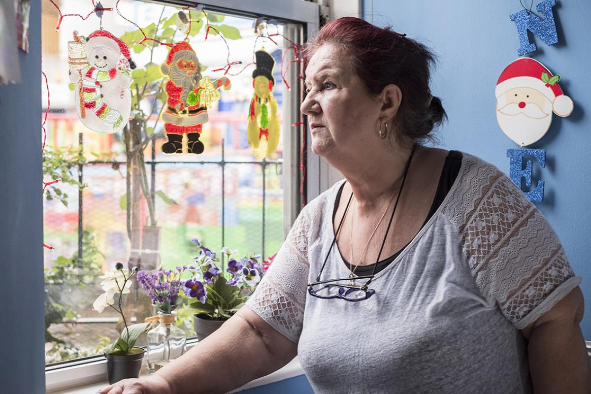 Betty is standing looking out of her living room window with her forearm resting on the sill. Christmas motifs hang from the window; a snowman, Santa Claus and a snowman with a black top hat. On the sill are two flower pots and a glass bottle of purple flowers. Hanging on the blue wall is the head of Santa Claus with a large letter N above and an E below. Betty is wearing a cream summer top with lacey short sleeves, has a thin gold chain necklace and glasses hanging from her neck.