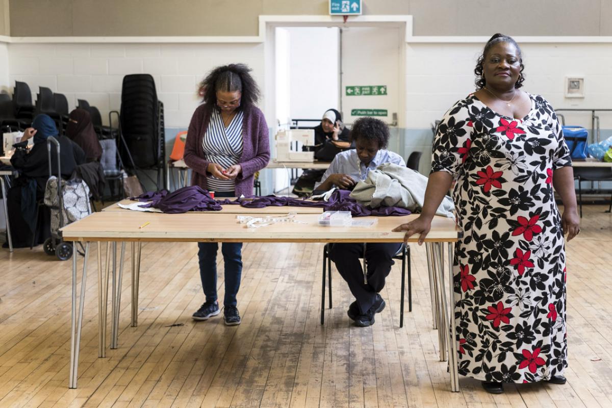 Esther is standing in a large wooden floored hall at Venture Centre with one hand leaning on a large table dressed in a floor length black and red flower print dress. Two other women are at the table, sewing - behind is another large table with a lady and a sewing machine. In the right hand corner is a lady at a table with a sewing machine and her shopping bag standing next to her.