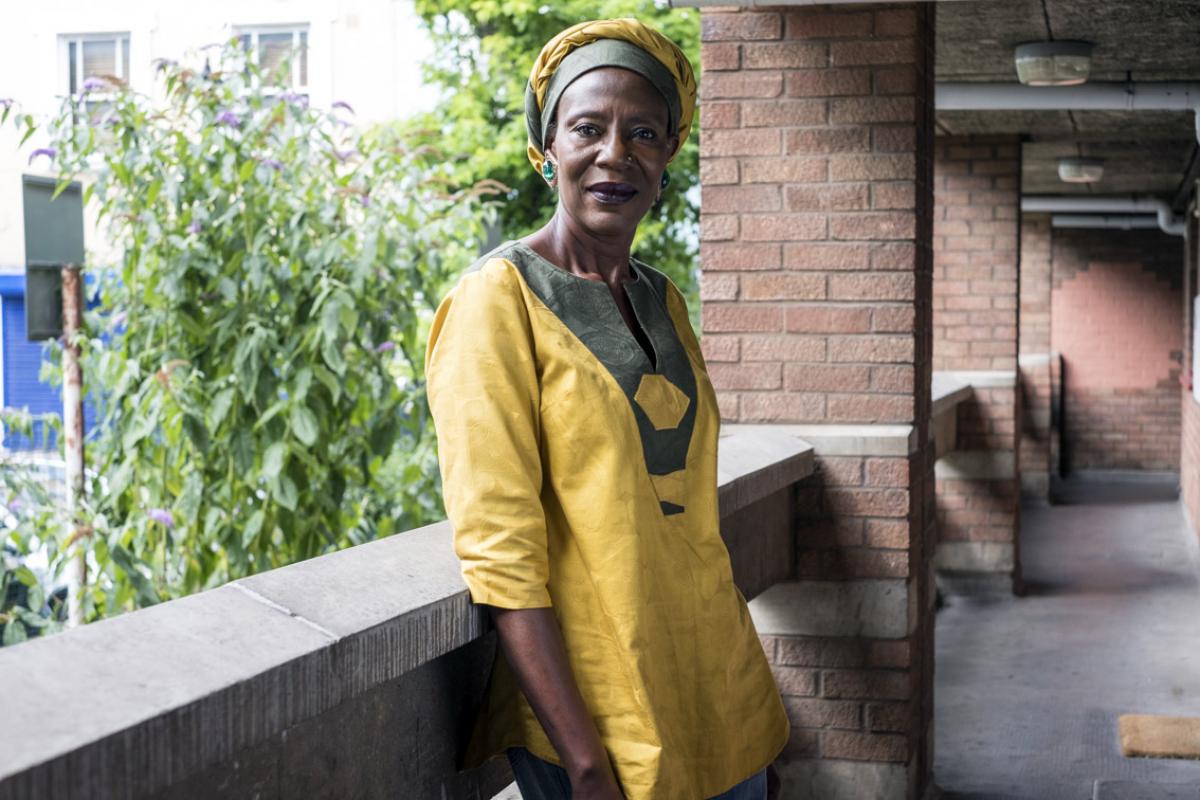 Grace is standing in one of the walkways that connects the flats. She is leaning against the wall that overlooks the trees showing in the background. Grace is wearing a yellow tunic with a dark green border around the neckline and a matching turban style hat.