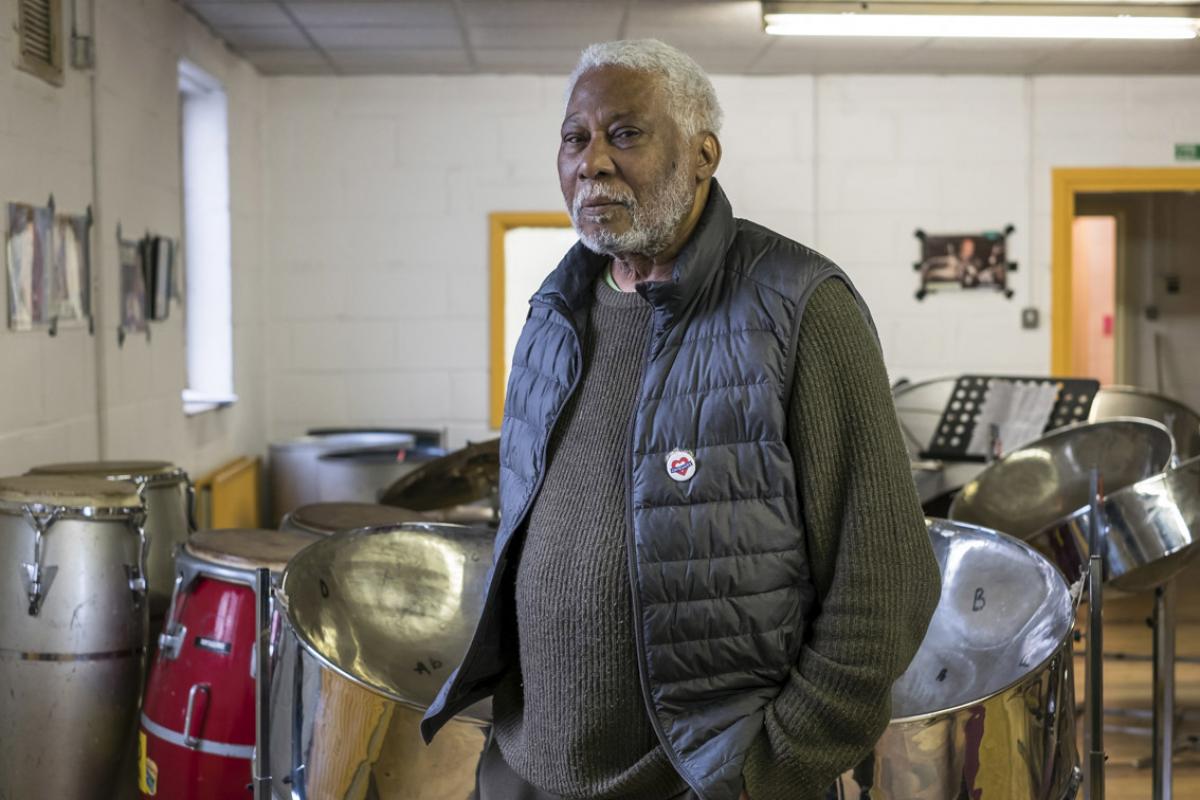 Leslie is standing in one of the rooms of the Venture Centre with several steel drums sitting in the background. The walls are painted white. Leslie is standing with his hands in his trouser pockets. He is wearing a dark grey jumper with a navy blue sleeveless puffer jacket over the top. He has short white hair and a short white moustache and beard.