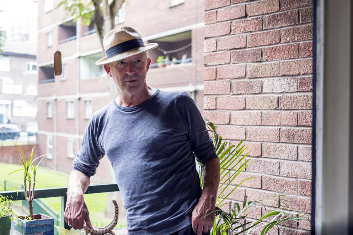 Micky is standing on the balcony with other Wornington Green Estate buildings in the background. Micky is leaning on the railing and is wearing a boater and a blue sweatshirt. There are plants to the left and right of him.