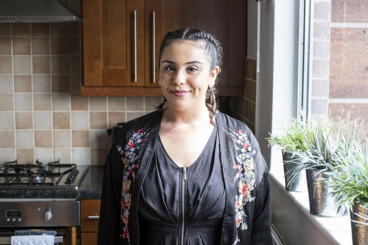 Oumayma is standing in her kichen. Behind her is a gas ring oven and a wooden cabinet on the wall, the tile surrounding are a mix of mushroom browns and cream. To her left there are three plants in silver pots. Oumayma in wearing a fitted black cotton top with a zip down the front and a black collarless jacket over the top that has flowers embroidered down either side of the front.