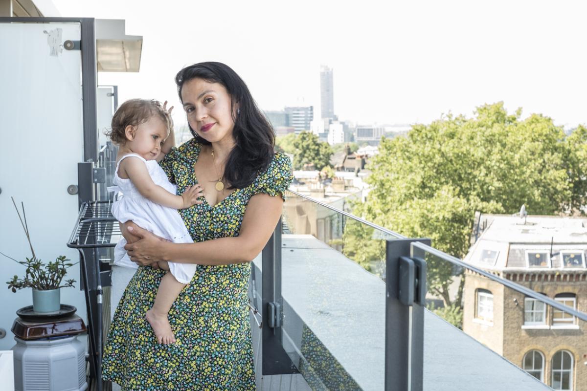 Vanessa is standing on the balcony of her new flat, in the background there are trees and buildings of various heights. Off to the left is a plantpot and just behind an empty clotheshorse. Vanessa is holding her little girl on her right hip who is wearing a white dress. Vanessa is wearing a summer dress with a small flowery print in yellow, blue and dusty pink.